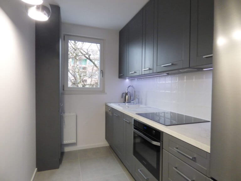 Zdjęcie przedstawia industrialną kuchnię na wymiar, która łączy w sobie funkcjonalność z nowoczesnym designem. Szafki w głębokim grafitowym kolorze zapewniają wyrafinowany wygląd, który jest zarówno nowoczesny, jak i ponadczasowy. Stonowana kolorystyka szafek kontrastuje z jasnym, delikatnym blatem, który wydaje się być inspirowany wzorem marmuru. Całość jest oświetlona za pomocą subtelnych świateł LED umieszczonych pod szafkami górnymi, co podkreśla czystość i linię płytek ściennych. Firma Aksent, jako doświadczony producent kuchni na wymiar, ma na uwadze, że sercem każdego domu jest kuchnia. Dlatego każdy projekt realizowany jest z największą starannością, mając na celu stworzenie nie tylko pięknej przestrzeni, ale przede wszystkim funkcjonalnej, która ułatwi codzienne czynności. Aksent rozumie, że kuchnia na wymiar to inwestycja, która ma służyć przez lata, dlatego wybiera materiały o wysokiej trwałości i proponuje inteligentne rozwiązania dopasowane do indywidualnego stylu życia klienta.