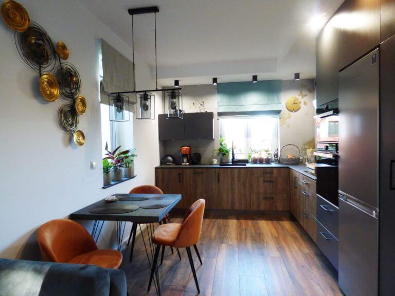 Zdjęcie przedstawia przestronną kuchnię, która łączy nowoczesność z klasycznymi elementami. Dominują tu ciepłe odcienie drewna widoczne w szafkach i na podłodze, które komponują się z eleganckimi, matowymi sprzętami AGD. Centralne miejsce zajmuje stół z ciemnego drewna, przy którym stoją cztery krzesła tapicerowane w odcieniu rustykalnego pomarańczu. Dodatkową dekoracją są złote, abstrakcyjne talerze na ścianie, nadając wnętrzu artystycznego charakteru. Przez okno wpada naturalne światło, które podkreśla zielone rośliny na parapecie. Kuchnia jest wykonana na zamówienie, co jest specjalnością firmy Aksent, producenta kuchni na wymiar. Taka personalizacja pozwala na doskonałe dopasowanie mebli do przestrzeni i potrzeb użytkownika. Oferując kompleksową usługę, od projektowania po montaż, firma Aksent prawdopodobnie dostarcza swoim klientom nie tylko meble, ale także profesjonalne doradztwo w zakresie ergonomii, stylu i funkcjonalności. Indywidualne podejście do każdego projektu zapewnia unikalność każdej kuchni, co czyni je idealnym rozwiązaniem dla osób poszukujących niepowtarzalnego wnętrza.