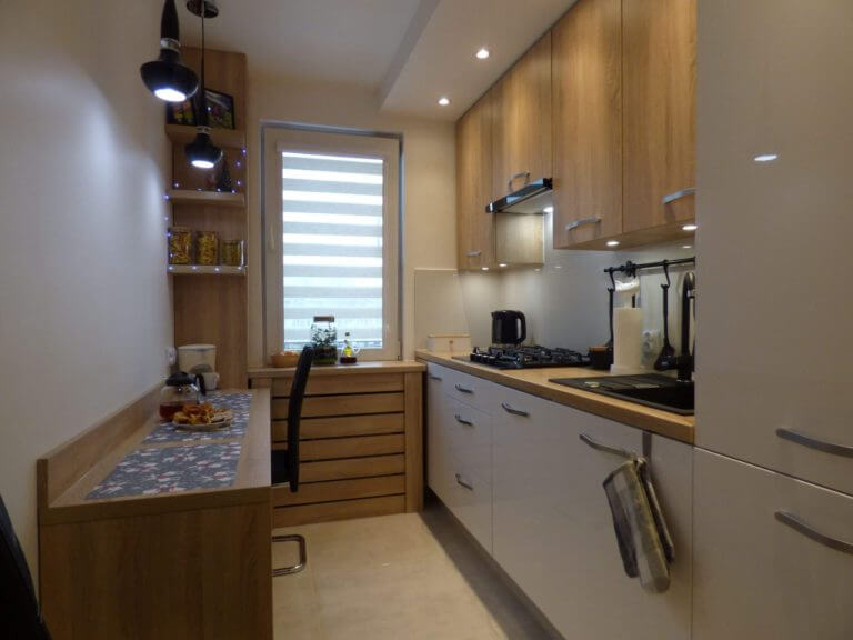 Na zdjęciu przedstawiona jest nowoczesna kuchnia wykonana na wymiar, w której dominuje połączenie bieli szafek i ciepłego koloru drewnianego blatu. Wnętrze jest jasne i przytulne, z dużym oknem, które wpuszcza naturalne światło, dodając przestrzeni ciepła i głębi. W kuchni tej znajdziemy również starannie dobrane akcesoria, które podkreślają jej funkcjonalny charakter. Firma Aksent, odpowiedzialna za stworzenie tej kuchni, specjalizuje się w projektowaniu i wykonawstwie mebli na wymiar, co pozwala na idealne dopasowanie do unikalnych wymiarów i potrzeb każdego klienta. Takie indywidualne podejście zapewnia maksymalne wykorzystanie dostępnej przestrzeni, a także możliwość stworzenia spersonalizowanej i jednocześnie optymalnie funkcjonalnej kuchni.