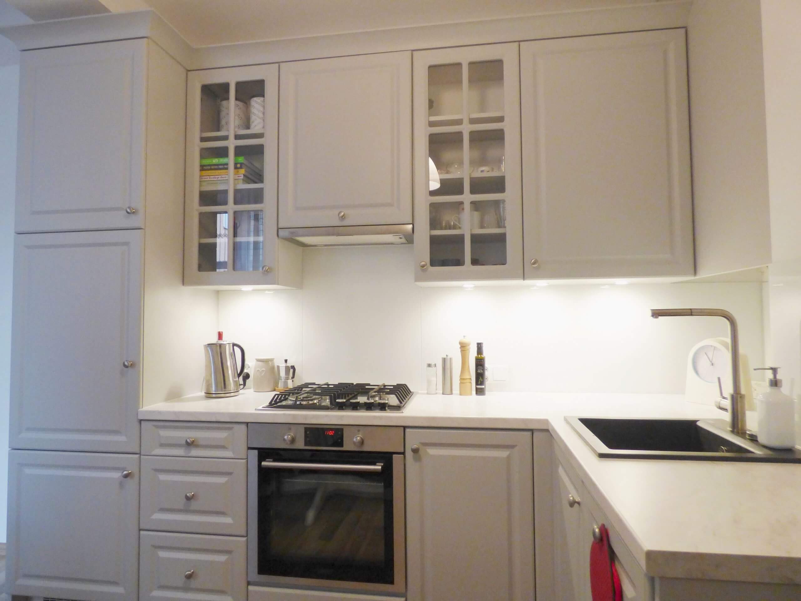 Na zdjęciu zaprezentowano elegancką kuchnię na wymiar, która charakteryzuje się subtelną kolorystyką i ciepłym, przyjemnym oświetleniem. Jasnobłękitne szafki z prostymi, ale eleganckimi uchwytami tworzą wyważony i spokojny nastrój. Kuchnia wyposażona jest w nowoczesne urządzenia AGD, w tym piekarnik i płytę gazową, co wskazuje na jej funkcjonalność. Szklane drzwiczki górnych szafek dodają lekkości, co zwiększa poczucie przestronności. Kuchnia jest zaprojektowana z myślą o maksymalnym wykorzystaniu przestrzeni, zachowując przy tym czystą i organizowaną estetykę. Jako producent kuchni na wymiar, firma Aksent rozumie potrzebę tworzenia przestrzeni, które są zarówno piękne, jak i praktyczne. Ta kuchnia jest odzwierciedleniem ich rzemiosła, w której zastosowano precyzyjnie wykonane meble, zaprojektowane tak, aby spełniać wymagania i pasować do konkretnego stylu życia klienta. Aksent skupia się na szczegółach, od wyboru materiałów, przez kolorystykę, po wybór akcesoriów i sprzętu AGD, aby każda kuchnia była nie tylko centrum domowej aktywności, ale również odzwierciedleniem osobistych preferencji użytkownika. Wizja firmy Aksent to tworzenie przestrzeni kuchennych, które łączą w sobie wyrafinowanie i funkcjonalność, dostosowane do indywidualnych wymagań każdego klienta.