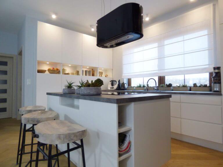 Zdjęcie przedstawia nowoczesną kuchnię na wymiar z centralnie umieszczoną wyspą kuchenną. Jasne, minimalistyczne szafki tworzą spójny i elegancki wygląd, który jest uzupełniony przez ciemny blat kontrastujący z białą kolorystyką mebli. Nad wyspą kuchenną zawieszony jest czarny, nowoczesny okap, który stanowi mocny akcent dekoracyjny. Trzy drewniane stołki barowe o nieregularnych kształtach siedzisk nadają wnętrzu odrobinę rustykalnego ciepła. Duże okno zapewnia obfitość naturalnego światła, co dodatkowo rozświetla przestrzeń, a rośliny doniczkowe na blacie dodają kuchni domowego charakteru. Wnętrze to perfekcyjne połączenie funkcjonalności i nowoczesnego designu. Firma Aksent zyskała uznanie jako producent kuchni na wymiar, łączący w sobie nowoczesne rozwiązania z indywidualnymi potrzebami klientów. Każdy projekt jest dopracowany do perfekcji, co sprawia, że nasze kuchnie są nie tylko wizualnie atrakcyjne, ale także ergonomiczne i wygodne w codziennym użytkowaniu. W Aksent wierzymy, że kuchnia jest sercem domu, dlatego do każdego zlecenia podchodzimy z niezwykłą starannością, aby spełnić oczekiwania nawet najbardziej wymagających klientów. Skontaktuj się z nami, abyśmy mogli stworzyć kuchnię, która będzie idealnie pasować do Twojego stylu życia i domowego wnętrza.