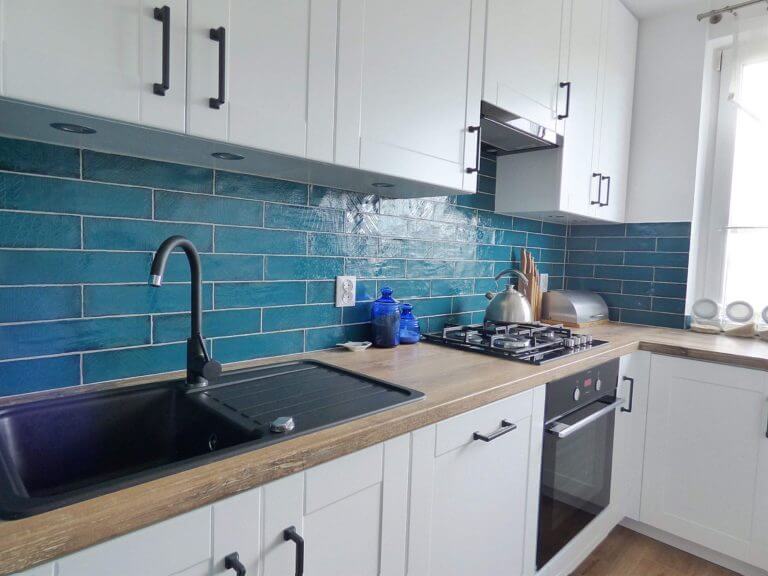 Zdjęcie przedstawia nowoczesną, jasną kuchnię z białymi szafkami i ciemnoniebieską, błyszczącą ścianą. Blaty są wykonane z jasnego drewna, a całe pomieszczenie jest dobrze oświetlone dzięki dużemu oknu. Widać czarny zlew z nowoczesnym kranem, a obok niego umieszczono kuchenkę z czterema palnikami i metalicznym czajnikiem. Na blacie obok kuchenki poukładano różne przybory kuchenne, w tym talerze i kubki. Firma Aksent to producent kuchni na wymiar, który doskonale rozumie indywidualne potrzeby swoich klientów. Każda kuchnia jest projektowana z myślą o funkcjonalności i estetyce, gwarantując najwyższą jakość wykonania i trwałość. Dzięki firmie Aksent Twoja kuchnia stanie się miejscem, gdzie gotowanie będzie prawdziwą przyjemnością. Firma oferuje szeroki wybór materiałów i wykończeń, co pozwala na stworzenie unikalnego wnętrza dostosowanego do stylu życia każdego klienta.