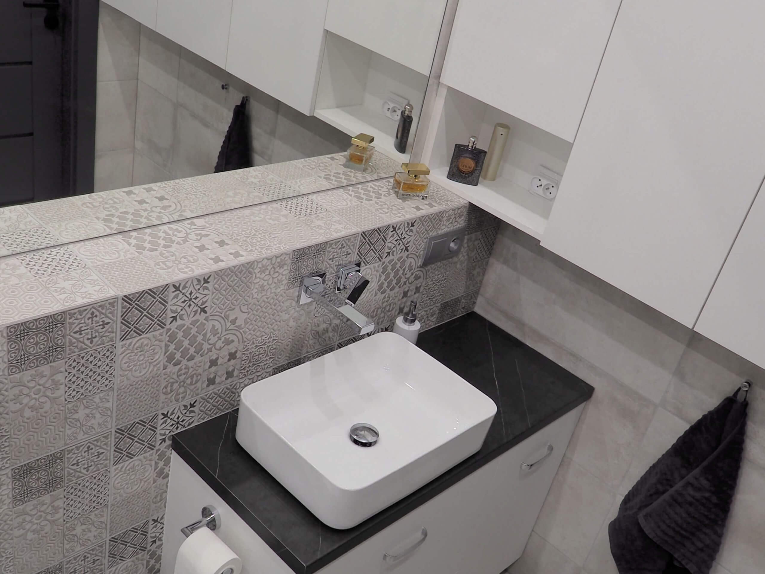 Na zdjęciu jest łazienka z nowoczesnym wystrojem. Centralnym elementem jest biała, prostokątna umywalka umieszczona na szafce w jasnym odcieniu z dwoma szufladami. Nad umywalką wisi lustro zamykające się szafką. Szafka umywalkowa i ściana zostały wykończone płytkami w dekoracyjne wzory w odcieniach szarości, które nadają wnętrzu elegancki, ale i współczesny charakter. Po prawej stronie widać włączniki oraz kosmetyki, co świadczy o funkcjonalności przestrzeni. Firma Aksent, jako producent mebli na wymiar, mogłaby być odpowiedzialna za wykonanie takiej szafki umywalkowej, zaprojektowanej na specjalne zamówienie, aby dopasować się do wymiarów i stylu łazienki. Produkty na wymiar są świetnym wyborem dla tych, którzy chcą maksymalnie wykorzystać przestrzeń i nadać jej indywidualny charakter. Aksent, specjalizując się w tego typu meblarstwie, oferuje możliwość stworzenia unikalnych rozwiązań, które są zarówno praktyczne, jak i estetycznie dopasowane do upodobań klienta.
