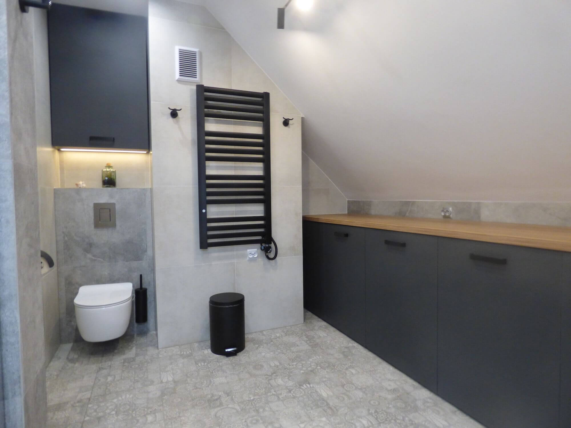 Na zdjęciu widać elegancko urządzoną łazienkę z elementami wykonanymi na wymiar. Design jest nowoczesny i funkcjonalny, a kolorystyka opiera się na spokojnej palecie szarości i czerni. Łazienka wyposażona jest w wiszącą toaletę. Przestrzeń jest dobrze zaplanowana z dużymi szarymi płytkami ceramicznymi na ścianie i pasującymi do nich szafkami łazienkowymi. Szafki są ciemne, z drewnianym blatem, dodającym ciepła wnętrzu. Uzupełnieniem aranżacji jest czarny grzejnik łazienkowy, który stanowi zarówno element grzewczy, jak i dekoracyjny. Aksent specjalizuje się w tworzeniu mebli łazienkowych na wymiar, które idealnie wpasowują się w potrzeby i wymogi każdej przestrzeni. Oferujemy meble, które łączą w sobie wytrzymałość i funkcjonalność, niezbędne w wilgotnych warunkach łazienki. Nasi klienci mogą wybierać spośród szerokiej gamy kolorów i materiałów, co pozwala na stworzenie spersonalizowanego wnętrza łazienki w zgodzie z ich własnym gustem i stylem życia. Aksent dba o to, by każdy mebel był nie tylko estetycznie atrakcyjny, ale również praktyczny, wykorzystując innowacyjne rozwiązania do przechowywania i organizacji przestrzeni. Meble łazienkowe Aksent są synonimem jakości, trwałości i designu na najwyższym poziomie.