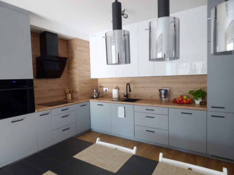 Na prezentowanym obrazku widzimy nowoczesną kuchnię, której projekt opiera się na eleganckiej kombinacji naturalnego drewna oraz szaro-białych frontów szafek. Kuchnia wyposażona jest w wysokiej klasy sprzęt AGD, co w połączeniu z wyrafinowanym oświetleniem i funkcjonalnym układem tworzy przemyślaną przestrzeń do gotowania i spożywania posiłków. Szafki i szuflady na wymiar zapewniają maksymalne wykorzystanie dostępnej przestrzeni, a ich wykończenie w stonowanych odcieniach wprowadza atmosferę spokoju i harmonii. Całość dopełniają eleganckie, wiszące lampy, które nadają wnętrzu nowoczesny charakter. Aksent specjalizuje się w tworzeniu mebli kuchennych na wymiar, które odpowiadają najnowszym trendom w designie i ergonomii. Nasza oferta obejmuje indywidualne projekty, które są dostosowane do specyficznych wymagań i oczekiwań naszych klientów. Wykorzystujemy materiały najwyższej jakości, a nasze meble są zaprojektowane tak, aby zapewnić trwałość, funkcjonalność i estetyczny wygląd. Dbamy o to, by każda kuchnia była nie tylko piękna i nowoczesna, ale także praktyczna i komfortowa w codziennym użytkowaniu. Z Aksent klient ma pewność, że jego kuchnia będzie miejscem, które inspiruje i sprawia radość z gotowania każdego dnia.
