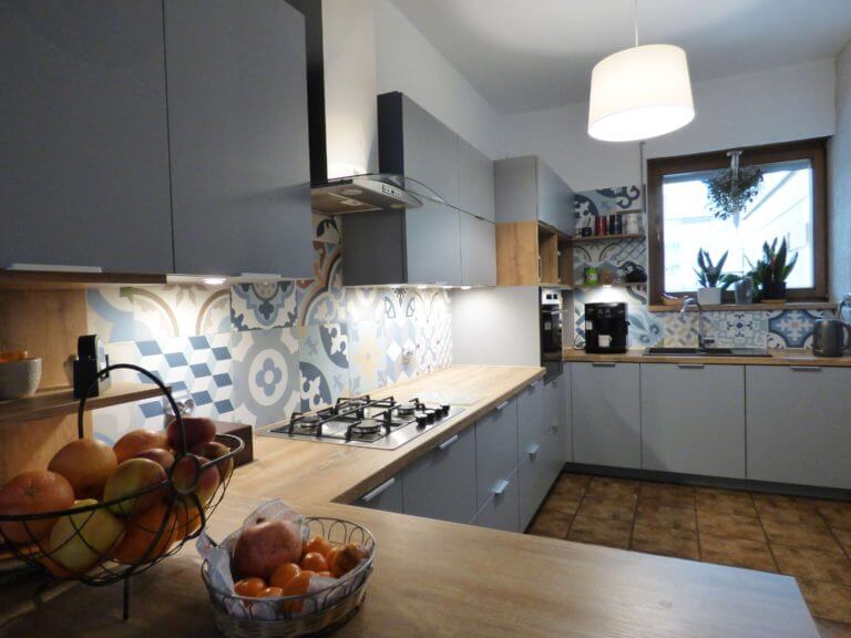 Na zdjęciu widoczna jest nowoczesna kuchnia na wymiar, charakteryzująca się szarymi frontami szafek oraz drewnianym blatem. Kuchnia jest dobrze oświetlona, z lampą wiszącą nad blatem. Posiada także ozdobne płytki ze wzorami w odcieniach niebieskiego, szarego i żółtego nad blatem roboczym, co dodaje jej unikalnego charakteru. Wyposażenie obejmuje zintegrowane urządzenia kuchenne oraz stojącą na blacie ekspres do kawy. Całość komponuje się w elegancki sposób, łącząc funkcjonalność z nowoczesnym designem. Oferta Aksent to idealne rozwiązanie dla osób, które cenią sobie wysoką jakość wykonania oraz indywidualne podejście do potrzeb klienta. Dzięki możliwości wykonania na wymiar, każdy element kuchni może być dostosowany do specyfiki pomieszczenia, co jest szczególnie ważne w przypadku nierównych ścian czy nietypowych układów pomieszczeń. Szare fronty to wybór idealny dla tych, którzy poszukują nowoczesnego, a zarazem ponadczasowego wyglądu swojej kuchni, natomiast drewniany blat wprowadza ciepły i przytulny akcent, tworząc przyjazną przestrzeń do gotowania i spędzania czasu z rodziną i przyjaciółmi.