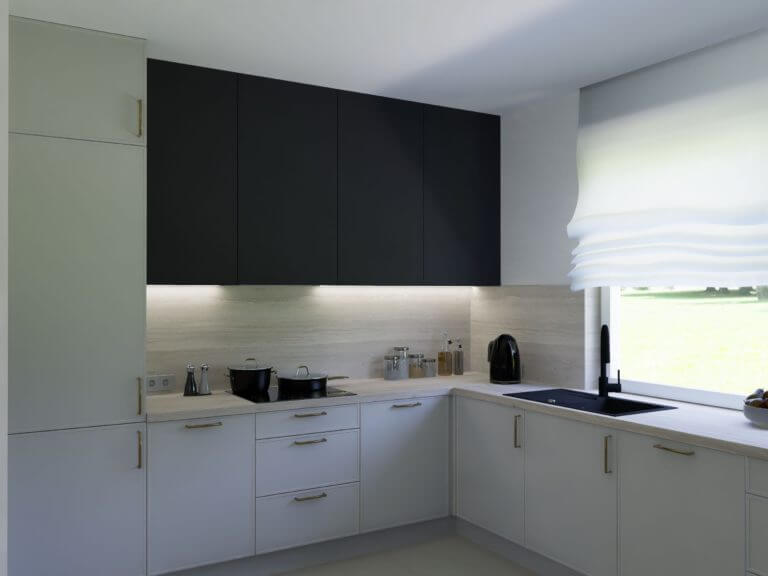 Na przedstawionym zdjęciu widzimy przestrzenną kuchnię na zamówienie, w której biel szafek stwarza jasne i czyste tło dla ciemnych, eleganckich górnych szaf. Proste, ale stylowe złote uchwyty doskonale komplementują kolorystykę, a jednolite, czarne wykończenie zlewozmywaka i baterii kuchennej podkreślają nowoczesny charakter pomieszczenia. Naturalne światło płynie przez okno, a uroczy, miękko falujący zasłon przepuszcza delikatne światło do wnętrza, tworząc spokojną i przytulną atmosferę. Firma Aksent, jako doświadczony producent kuchni na wymiar, specjalizuje się w projektowaniu takich przestrzeni, które łączą indywidualne potrzeby klienta z najnowszymi trendami w projektowaniu wnętrz. Kuchnie wykonane przez Aksent charakteryzują się nie tylko wyjątkowym designem, ale również wysoką funkcjonalnością i ergonomią. Dzięki rzemieślniczej precyzji i zastosowaniu materiałów wysokiej jakości, każda kuchnia jest dostosowana do specyficznych wymagań, oferując trwałość i wygodę użytkowania na najwyższym poziomie. Personalizacja idzie w parze z perfekcją wykonania, co sprawia, że każda kuchnia na zamówienie od Aksent jest wyjątkowym sercem domu, odpowiadającym na dynamiczne życie współczesnych użytkowników.