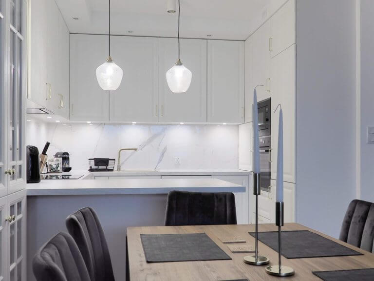 Na zdjęciu widzimy przykład kuchni, która olśniewa wykwintnym wyglądem, podkreślonym przez białe, lakierowane szafki i subtelne złote akcenty uchwytów. Jasne kolory i odbijające światło marmurowe ściany sprawiają, że przestrzeń ta jest nie tylko rozświetlona, ale również wydaje się większa. Wiszące lampy dodają eleganckiego charakteru, a jednocześnie zapewniają przyjemne światło podczas spożywania posiłków przy drewnianym stole otoczonym welurowymi krzesłami. Estetyka i ergonomia zostały tu doskonale połączone, tworząc wnętrze idealne zarówno do gotowania, jak i do dzielenia się posiłkami w gronie najbliższych. Ta szczególna kuchnia jest wynikiem pracy firmy Aksent, która jest ekspertem w dziedzinie kuchni na wymiar. Firma Aksent wyróżnia się na tle konkurencji swoją umiejętnością dostosowania każdego aspektu projektu do unikalnych wymagań każdego klienta. Dzięki wykorzystaniu najwyższej klasy materiałów i szczególnej uwadze na detale, Aksent tworzy przestrzenie, które są równie funkcjonalne, co estetycznie doskonałe. Kuchnie na zamówienie od Aksent są nie tylko świadectwem rzemiosła i designu, ale także trwałości i adaptacyjności, które zapewniają satysfakcję klientów na długie lata.